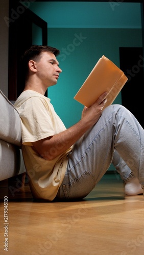 Fotografie Man in casual clothing sitting on a wooden floor, leaning against a sofa and eng