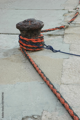 Fotografie An old rusty iron mooring bollard is wrapped with heavy orange and black ropes on a traditional stone pier