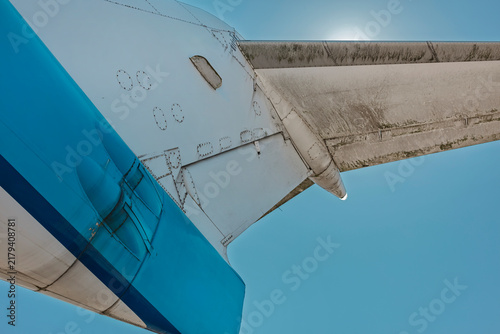 Картината върху платно Low angle detail of a passenger jet tail fin and stabilizer against the sun, showing weathered panels and a bold blue livery stripe
