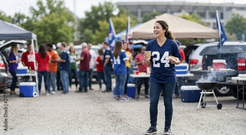 Smiling female football fan holding hot dog and drink at tailgate party. Woman in team jersey enjoying game day celebration in stadium parking lot