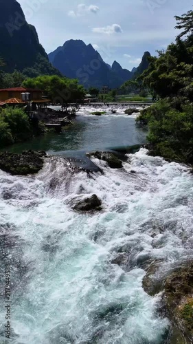 Vertical Video: Drone flying forward along a turquoise river valley with cascading white waterfall over a stone dam and karst mountains ahead
