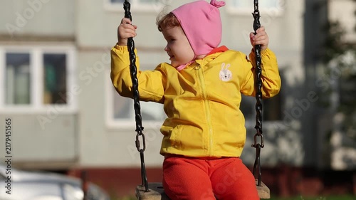 Little girl in pink hat and yellow jacket swinging on swing at playground. Happy child playing outdoors in autumn. Childhood, joy and family leisure activity concept
