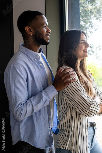 Foto Smiling interracial couple standing by a window in their modern home, looking ou