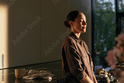 A contemplative woman stands in a sunlit modern kitchen, exuding calm and peace as she momentarily closes her eyes while connecting with her serene surroundings and mindful routines.