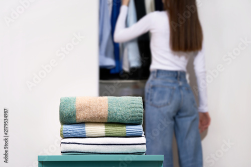 фотографія Folded woolen sweaters, pastel color stripes on blue drawer with woman near open wardrobe on light gray background
