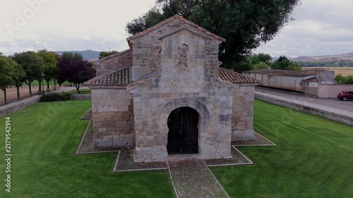 Aerial point-of-interest semicircular tracking shot highlighting stone masonry, horseshoe arch and facade of Visigothic church of San Juan de Baños under cloudy sky.