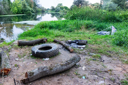 Old rubber tire and scattered logs arranged as a primitive makeshift fireplace seating area in a messy overgrown riverside spot with litter nearby, showing nature pollution and camping aftermath