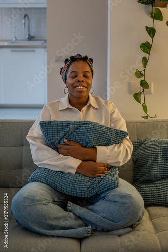 Konstfotografi Smiling african american woman embraces soft cushion while sitting cross-legged on sofa, daydreaming, expressing comfort, emotional warmth and contentment during serene self-care moment in cozy home