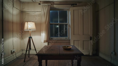 Old empty room at night with glowing tripod lamp, wooden table, bowl and window with curtain, dark hallway appears behind open door. Haunted house mystery and horror atmosphere concept