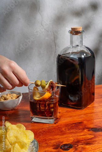 Bild auf Leinwand Hand holding gilda on a glass of vermouth on the rocks above a wooden table with