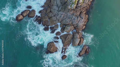Aerial drone view of turquoise ocean waves crashing against rugged coastal rocks in New Zealand.