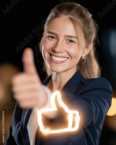 Woman Giving Thumbs Up: A smiling young woman, dressed in a business suit, gives a confident thumbs-up gesture, her face illuminated by the glow of a digital thumb icon.