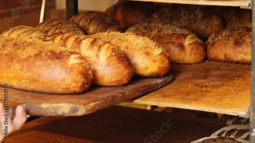 Baker transfers freshly baked bread from hot oven to wooden cooling rack. Skilled bakery worker uses sturdy wooden paddle to move loaves