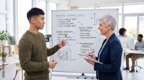 Colleagues planning year-end giving campaign on whiteboard in modern office. Nonprofit fundraising strategy meeting between young man and mature businesswoman
