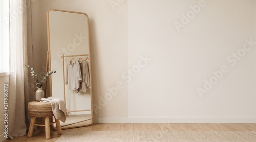 Minimal dressing room interior with floor mirror, wooden stool, hanging clothes, beige wall and wood floor, soft window light, empty space for fashion mockup and decor design.