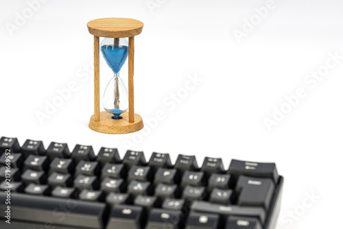 A wooden hourglass with blue sand nearly run out stands behind a black computer keyboard on a white background, symbolizing time, deadlines, and the consequences of pressing enter