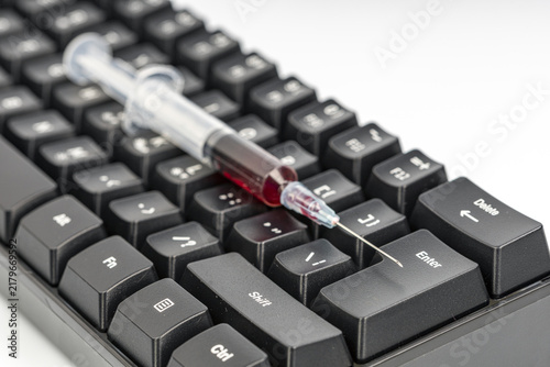Close-up shot of a medical syringe filled with red liquid resting near the enter key of a black computer keyboard, conceptually representing the health risks and dangers linked to online activity