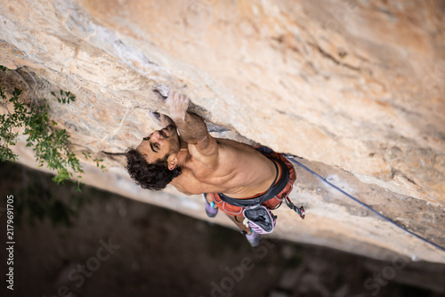 Shirtless climber grips a rocky overhang. His hands dig into the stone surface. He appears concentrated and determined. Climbing harness secures him to a rope. Green foliage peeks from the corner
