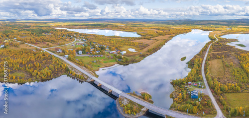 Fotografie Aerial panorama of Nedre Soppero village and the Muonio River bridge, with vibrant golden autumn forest and mirror-like water reflecting a dramatic cloudy sky