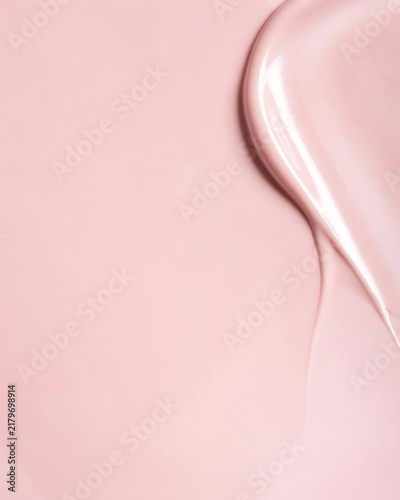 Close up of a smooth pink cosmetic cream texture with a curved swirl pattern and soft lighting on a plain light pink background surface.