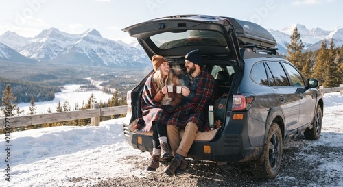 Happy young couple sitting in open car trunk drinking hot beverages during winter mountain road trip