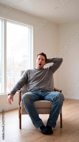 Man dozes and yawns in armchair stretching arm beside bright window light