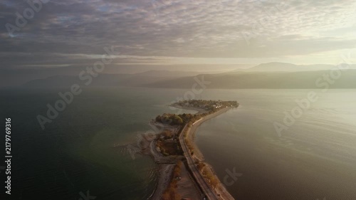 Long narrow causeway stretching into misty horizon and calm sea water on both sides filmed by drone. Soft dawn light and leading lines form minimal composition. Serene atmosphere evoking solitude 