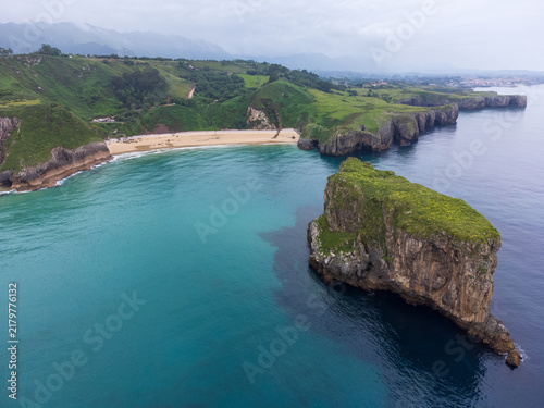 Aerial coastal view from the Atlantic Ocean overlooking calm waters, a rugged rocky islet, Playa de Andrín beach, lush green headlands, and cloud-covered mountains. Costa Verde, Asturias, Spain.