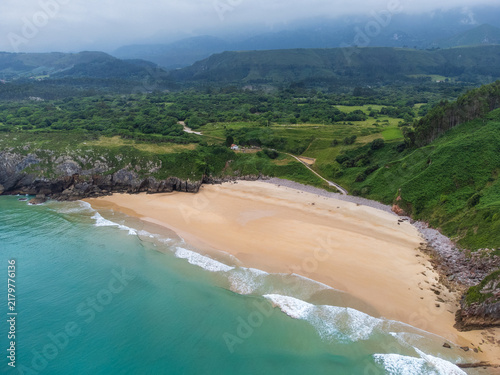 Aerial view from the sea toward the golden sands of Playa de Andrín beach, featuring coastal rock formations and green mountain ridges disappearing into cloud cover. Costa Verde, Asturias, Spain.