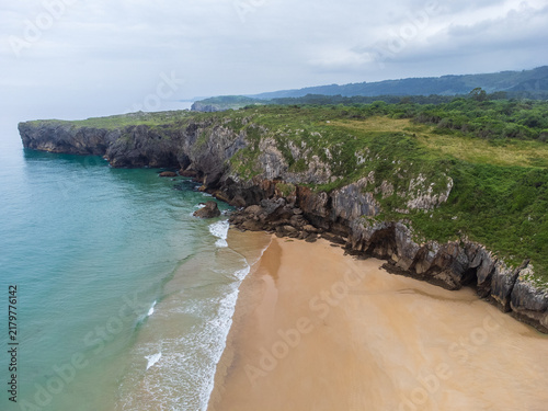 Aerial view of the empty sandy shores of Playa de Andrín, bordered by steep green cliffs stretching into the distance, with the calm Atlantic Ocean and distant mountains. Costa Verde, Asturias, Spain.