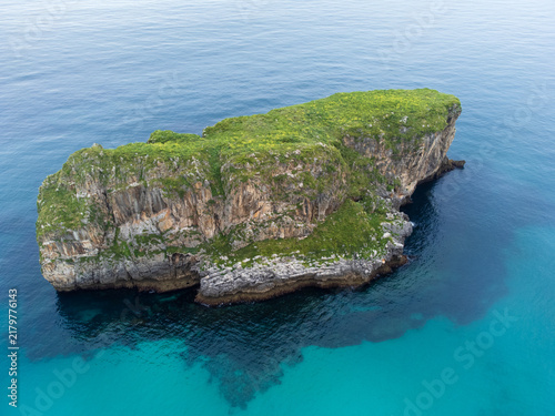 Minimalist aerial view of a solitary vegetation-covered rocky islet surrounded by the calm, turquoise waters of the Atlantic Ocean, Costa Verde, Asturias, Spain.