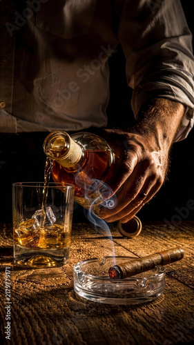 man pouring whiskey into glass with ice cubes on rustic table