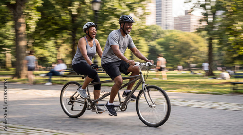 Couples Tandem Ride in Park: A joyous couple enjoys a tandem bicycle ride through a sun-dappled park, experiencing shared adventure and a healthy, active lifestyle amidst urban greenery.