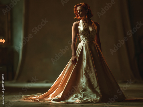 Moody low‑key portrait of short red‑haired woman in elegant strapless gold satin gown, dramatic soft side lighting against dark textured wall.