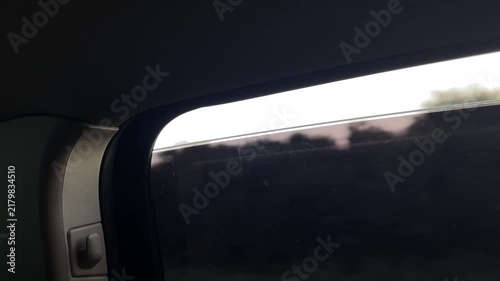 interior passenger corner view of tinted car window showing curved black trim, latch, textured headliner, faint glass smudges, and soft passing landscape blur window