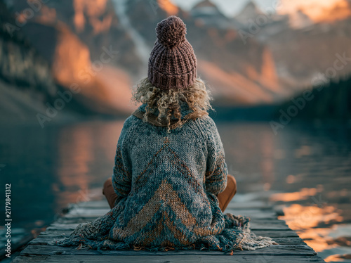 Woman sits cross‑legged meditating on a wooden dock beside calm water, warm sunset golden light glowing over peaceful lakeside scenery.
