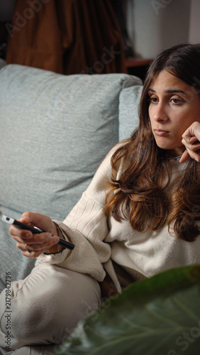 Cuadro en lienzo Young Latina woman sitting bored on a sofa while watching television, capturing idle time, home entertainment, media consumption, and everyday leisure