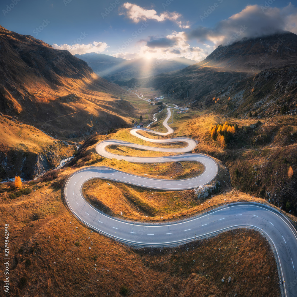 Obraz premium Aerial view of winding mountain road in the Swiss Alps, Switzerland, with sharp curves, golden slopes, valley and dramatic sun rays breaking through clouds over alpine peaks. Top drone view of road