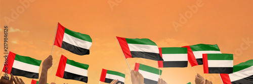 Fotografie Hands wave UAE flags against an orange sky during a celebration.