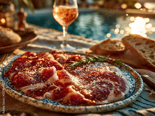 A romantic outdoor charcuterie spread by shimmering water, featuring cured meat, fresh bread, rose wine and glowing warm bokeh lights.