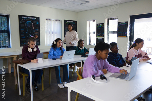Diverse youth working on laptops and tablet in classroom with headphones, instructor observing