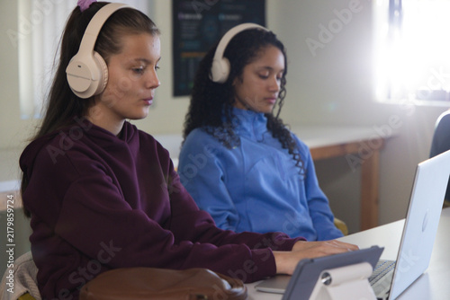 Two teen girls wearing over-ear headphones, studying on silver laptop and tablet at study table