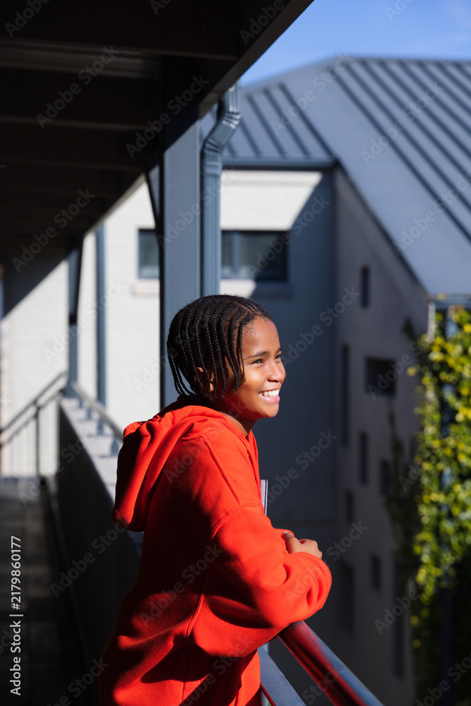 Obraz premium African American youth male wearing red hoodie leaning on balcony railing, smiling, looking right