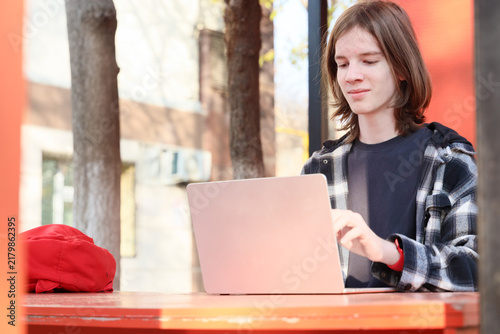 Young Student Works On Laptop Outdoors, Wearing Casual Plaid Shirt, With Focused Expression, Soft Sunlight, Blurred Urban Background, Useful For Education, Technology