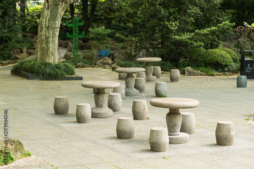 Photography Chinese park rest area with round stone tables and drum-shaped granite stools on a paved courtyard surrounded by green trees, Shanghai