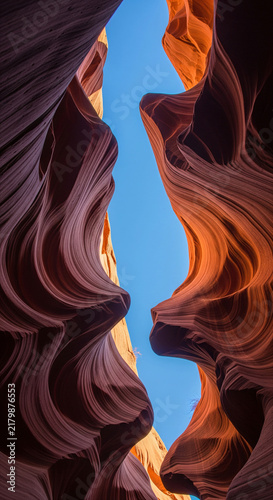 Stunning low angle view looking up through a narrow slot canyon with smooth, wavy red and orange sandstone walls against a clear blue sky. Abstract desert nature and geological rock formation.