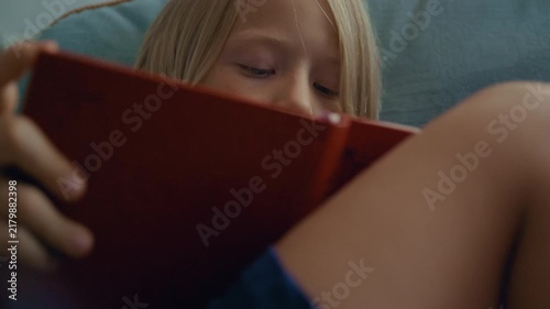Young child with long blond hair concentrating on reading a book with a red cover while resting