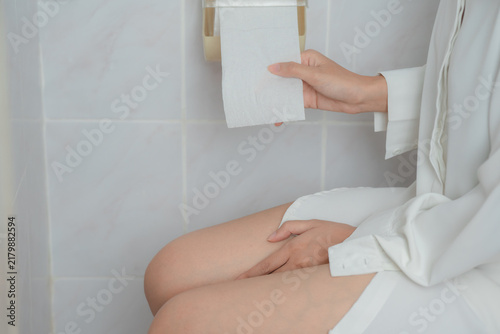Fotografie Constipation and digestive health concept showing a woman sitting on a toilet holding tissue in a bathroom, suggesting bowel difficulty