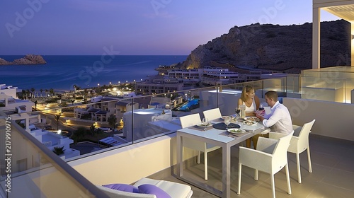 Couple dines on a balcony overlooking coastal city at dusk, with sea and mountains