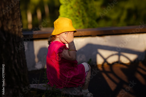 Little girl in yellow hat covering her face with hand while sitting outside. Child playing hide and seek in park. Portrait of shy kid outdoors. Summer recreation and childhood concept.